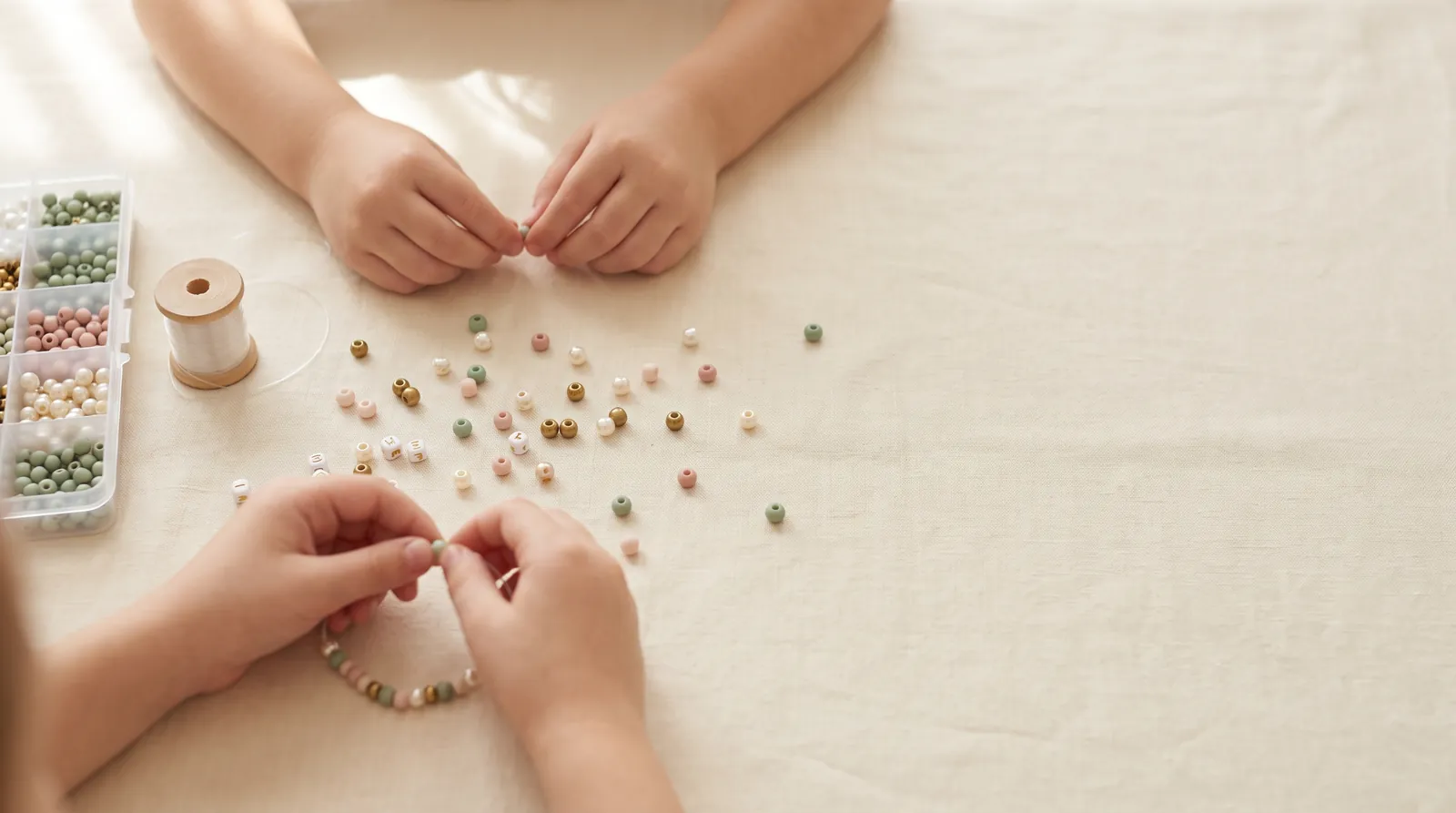 Hands stringing pastel and gold beads onto elastic cord on a cream linen table